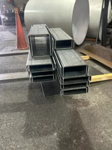 A stack of rectangular metal profiles rests on a floor, with a cone and rolled cylinders in the background.