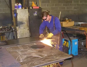 A metal fabricator in an orange-and-blue uniform operating a press brake to bend a metal sheet in a factory.