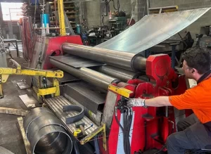A worker in an orange shirt operates a red plate rolling machine with four large rollers to curve a metal sheet.